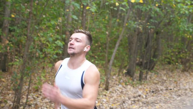 Athletic guy with wireless headphones sprinting fast along trail near forest. Young strong sportsman jogging outdoor at early autumn. Handsome man running at nature. Healthy active lifestyle concept