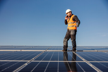 The worker is standing on the rooftop covered with solar panels and talking on the phone.