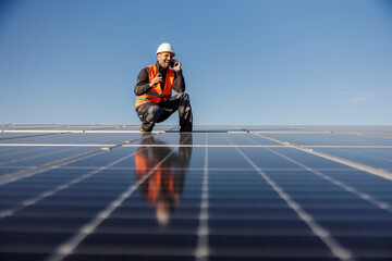 A happy worker is crouching next to the solar panels and talking on the phone.