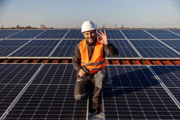 A happy worker with drill in his hands is showing okay gesture while couching next to the solar panels.