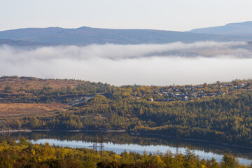 Foggy rural landscape. View of the village among the hills. In the background, fog in the mountains. Reservoir in the foreground. Magadan region, Siberia, Russian Far East. Autumn season, September.