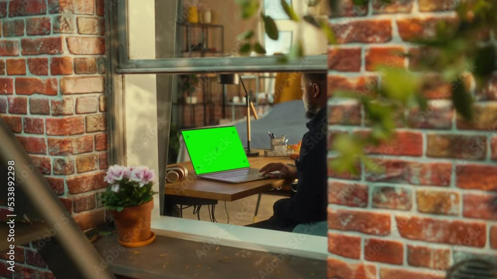 Aufkleber Black Man Using Laptop Computer with Green Screen Mock Up Display while Sitting Behind a Table in Stylish Living Room. Specialist Working on Computer, Browsing Internet. Shot from Outside the Window.