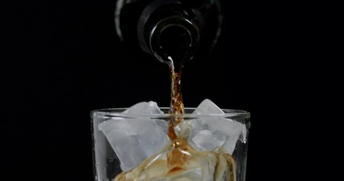 Slow motion shot of pouring cola from a bottle into a glass with ice cube.
