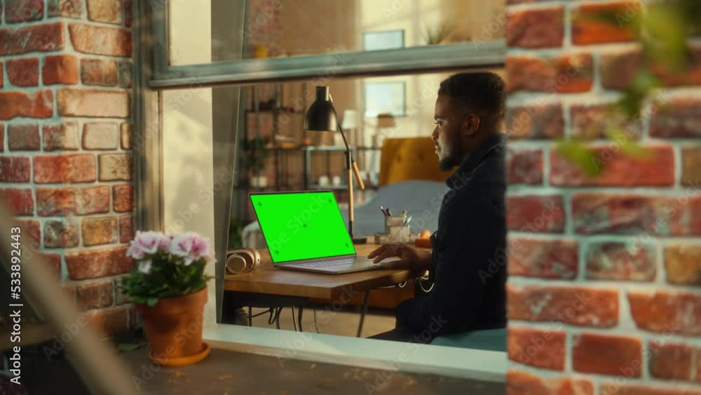 Poster African Man Working on a Laptop Computer with Green Screen Mock Up Display while Sitting at a Table in Stylish Living Room. Business Manager Working Remotely from Home. View from Outside the Window.