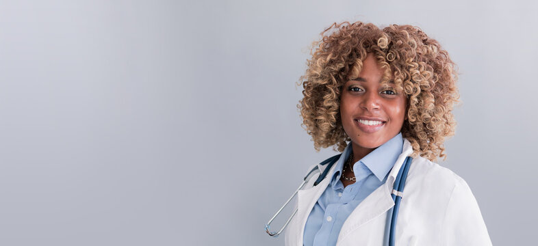 Smiling mixed race doctor in a and white coat, holding a stethoscope on a gray background looking at the camera, baner. Concept: doctor, medicine, practice, treatment of diseases
