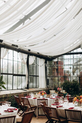 decorated greenhouse hall for a festive event with white capes and country chairs. numbered and decorated tables in a room with many windows and no people.	