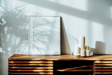 Modern minimalist Scandinavian style interior with white poster mockup and candles on a wooden console under sunlight and home plants shadows on a white gray wall. Living room design. Selective focus.