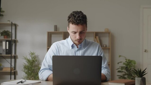 Brooding serious young man sit at table in comfortable home office room work on laptop
