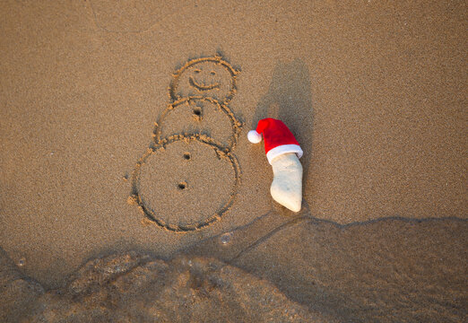 red Santa Claus hat on a white stone and a snowman painted on sand on coastal beach. The concept of celebrating Christmas holidays while traveling to warm tropical countries, New Year on the beach