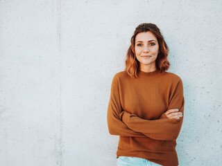 Woman in sweater with folded arms leans against concrete wall and looks to the side