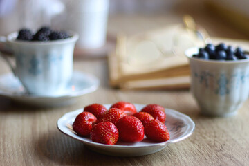 Vintage tea set filled with blueberries, blackberries and starwberries, open book, reading glasses and decorative candles on the table. Selective focus.