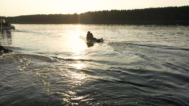 surfer lies on surfboard behind boat with sunset