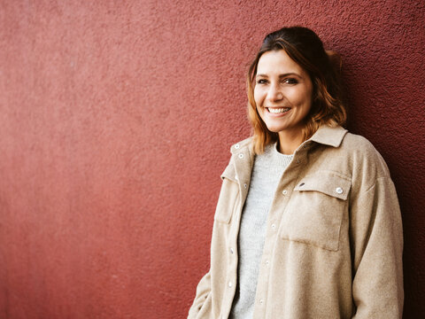 Woman stands in front of red wall and looks to the side laughing, copy space