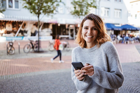 Young woman with cell phone stands in downtown and looks up at camera