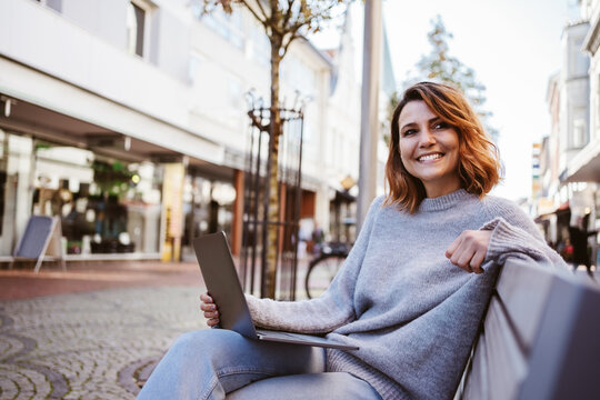 Laughing young woman with laptop sitting on park bench in downtown and looking to the side