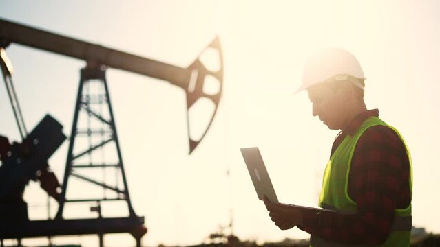 oil business. a worker works next to an oil pump holding a laptop. industry business oil and gas concept. engineer studying the level of oil lifestyle production on a laptop silhouette at sunset