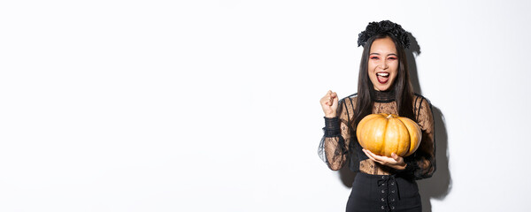 Image of excited beautiful asian woman celebrating halloween, wearing witch outfit and holding...