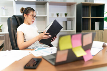 Fototapeta na wymiar asian beautiful woman focus on business work. overtime glasses female worker looking at file for document to done work. workplace full with sheet paper and sticky notes by motivation occupation girl