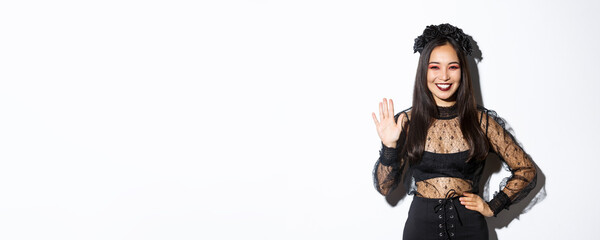 Image of friendly stylish asian woman in gothic lace dress waving hand to say hello, greeting...