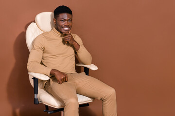 Portrait of attractive cheerful guy sitting in chair copy empty blank space ad isolated over brown...