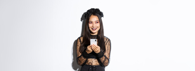 Portrait of cunning beautiful asian woman, witch in gothic lace dress using mobile phone, smiling...