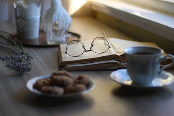 Cup of tea, plate with chocolate chip cookies, open book, reading glasses, lit candles and dry...