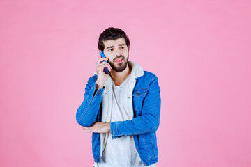 Young man in casual blue jacket and white shirt holding a smartphone against a pink background, capturing a selfie or taking a photo, with a modern and vibrant aesthetic
