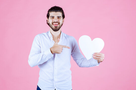 A person holding a white heart-shaped object against a pink background, dressed in a white shirt, pointing towards the heart with their right hand, conveying love or affection.