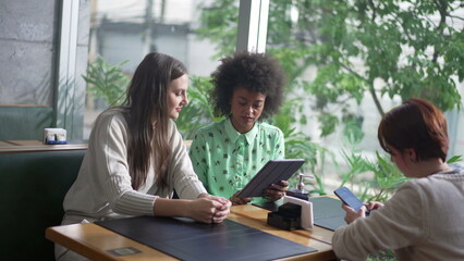 Woman showing tablet screen to female friend at coffee shop table. Three women interaction using modern technology devices