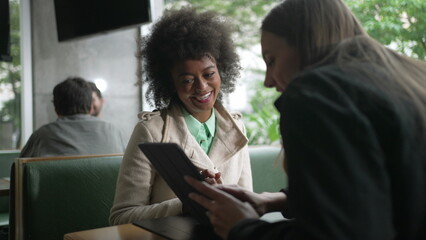 Two diverse women looking at tablet device at coffee shop. Woman sharing screen to friend sitting at cafe restaurant