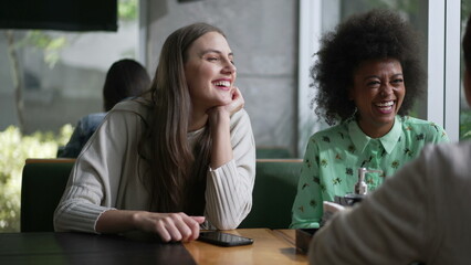 Three young diverse women laughing and smiling sitting at coffee shop table. Interaction between girlfriends in conversation. Real life laugh and smile