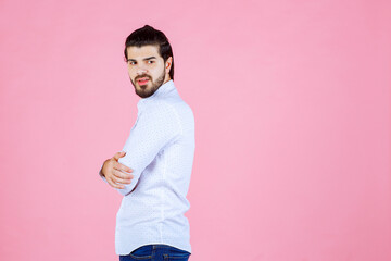 A person with short dark hair and light skin wearing a white long-sleeve shirt and jeans, standing against a solid pink background, with arms crossed and facing to the side
