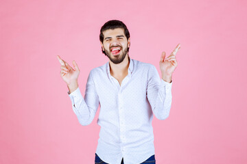 A person posing with peace signs in front of a pink background, wearing a white shirt and a light-colored blazer, exuding confidence and a cheerful demeanor