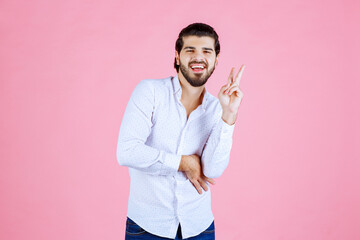 A person with a blurred face making a peace sign gesture against a solid pink background, dressed in a white shirt and dark pants, conveying a casual and friendly mood