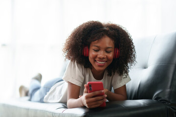 Bright and cute Asian young woman wearing headphones sit on the sofa and using smartphone to listen...