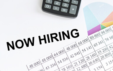 Now Hiring writing on a notebook next to reports and charts on a desk, a business concept