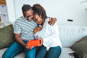 Young couple is at home. They are sitting on a couch, head to head and holding a present. Cheerful young woman receiving a gift from her boyfriend. Shot of a loving husband giving his wife a gift.