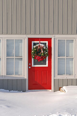 Fototapeta premium Vertical Christmas themed winter image of a vibrant red front door in a grey wooden building with a Christmas wreath hanging on the door dusted with freshly fallen snow.