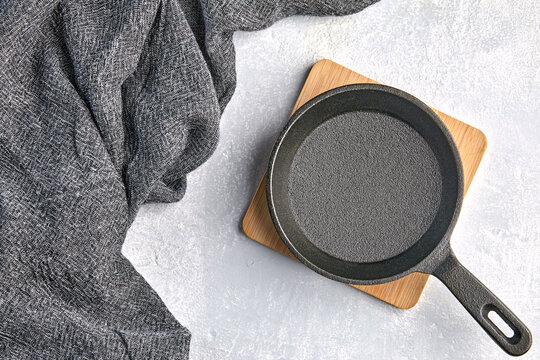 Black cast iron empty frying pan for one person on a wooden stand and a gray kitchen towel on a gray concrete background