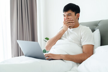 Sleepy man yawning on bed using laptop computer.