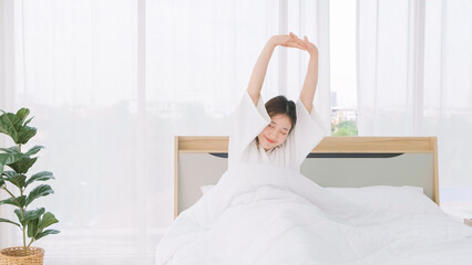 Fototapeta na wymiar Asian woman wearing bathrobe stretching her arms above her head while lying on bed. Morning relaxation routine after waking up. Woman tourist stretching her arms after waking up in bed of hotel room.