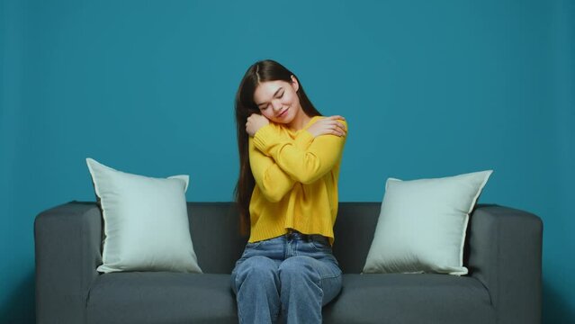 Smiling young girl hugs herself feeling self love and care, sitting on sofa. Body positive, acceptance, good self esteem