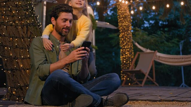Caucasian bearded man 30s father sitting at campsite in forest camping vacation near van trailer in evening looking smartphone mobile phone child daughter running to daddy embrace hugging dad