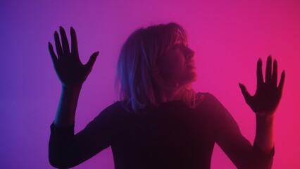 Frightened young woman touches glass with palms. Blonde examines glass and slides her hands over it with upset and afraid. Model poses in dark studio with pink purple lights and smoke. Close up.