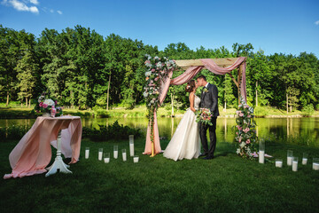 A couple of newlyweds during a wedding ceremony on the street near a decorated arch with flowers against the backdrop of a lake in the forest