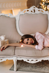 An attractive young woman with long hair in pink pajamas relaxes and sits on the sofa in a decorated room with a Christmas tree on Christmas holiday at home in the New Year. selective focus