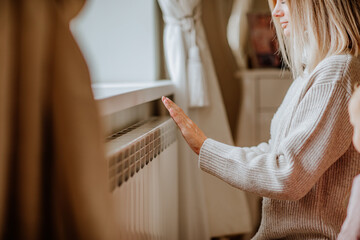 Young woman in long winter beige sweater is posing at home near the radiator - obrazy, fototapety, plakaty