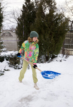 positive teenage girl in bright warm sportswear, clears the sidewalk, road, yard from snow after a strong snowstorm, discards the snow with a large shovel. Winter season, bad weather, cold, snow