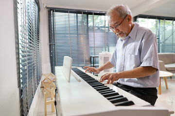 Happy smiling asian senior man with beard sitting and playing piano and singing a song in living...
