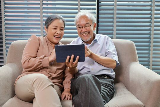 Happy smiling asian senior couple sitting on sofa and using tablet while online video call with friend or relative cousin at home living room. Internet information technology and lifestyle concept.